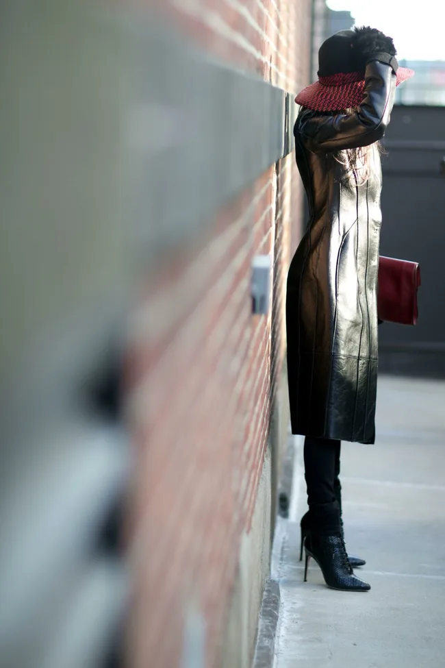 Person in long leather coat, red hat, black high-heeled boots, leaning against a brick wall.