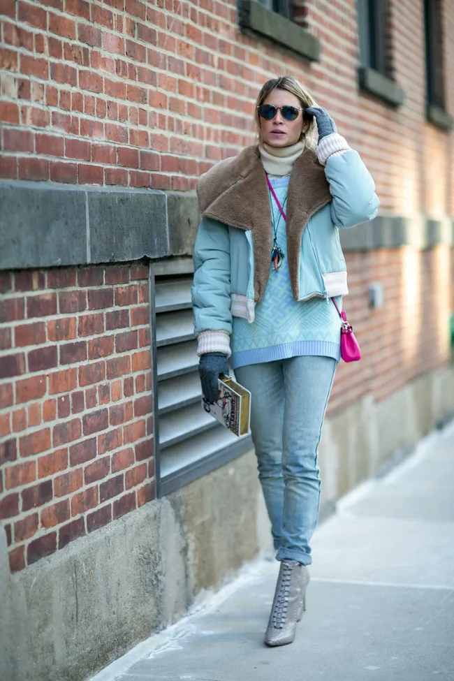 Woman in winter attire with a fur-lined jacket and blue jeans, walking along a brick wall, holding a book, wearing sunglasses.