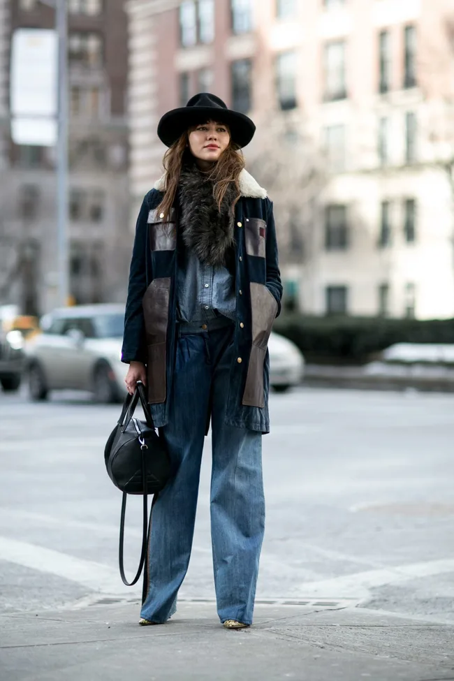 A woman stands at a city intersection in a wide-brim hat, denim outfit, long coat, and holding a black bag.