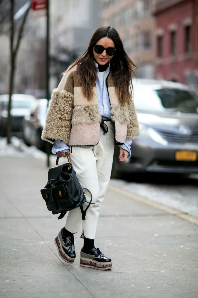 A woman in a stylish shearling jacket, white pants, and platform shoes walks down a city sidewalk carrying a black backpack.
