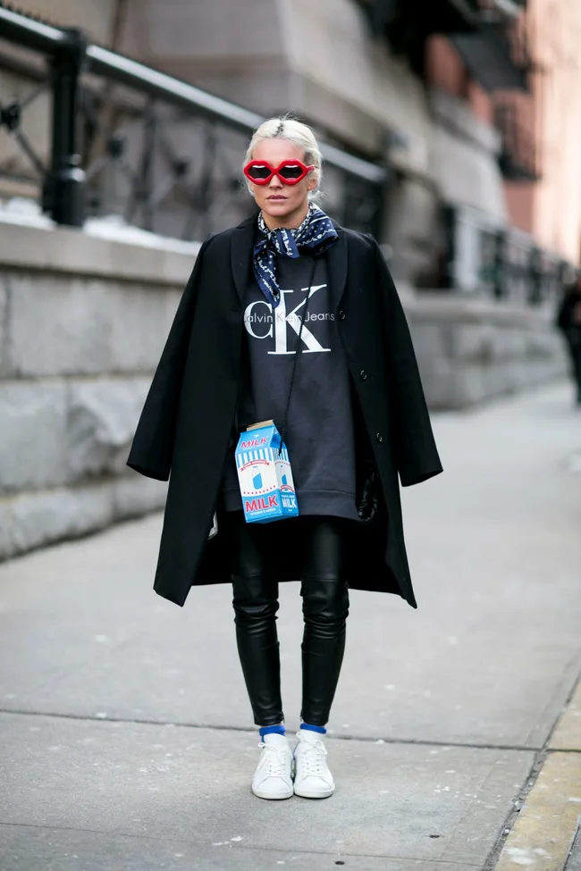 Blonde woman in red heart glasses, CK jacket, leather pants, white sneakers, with a milk carton bag on a city street.