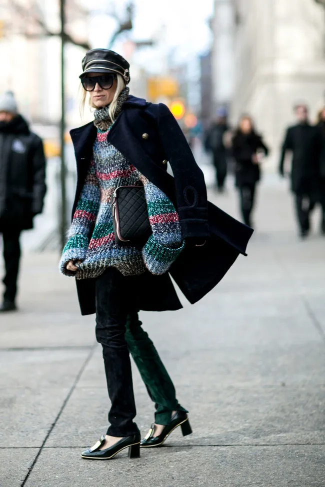 Street-styled person in a colorful sweater, black coat, jeans, heeled shoes, hat, and sunglasses with a quilted bag.