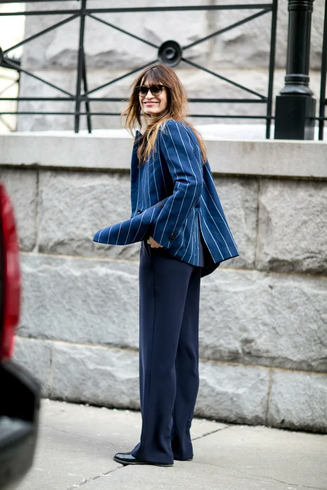 Woman in a blue pinstripe blazer, sunglasses, and navy pants, smiling outdoors near a stone wall.