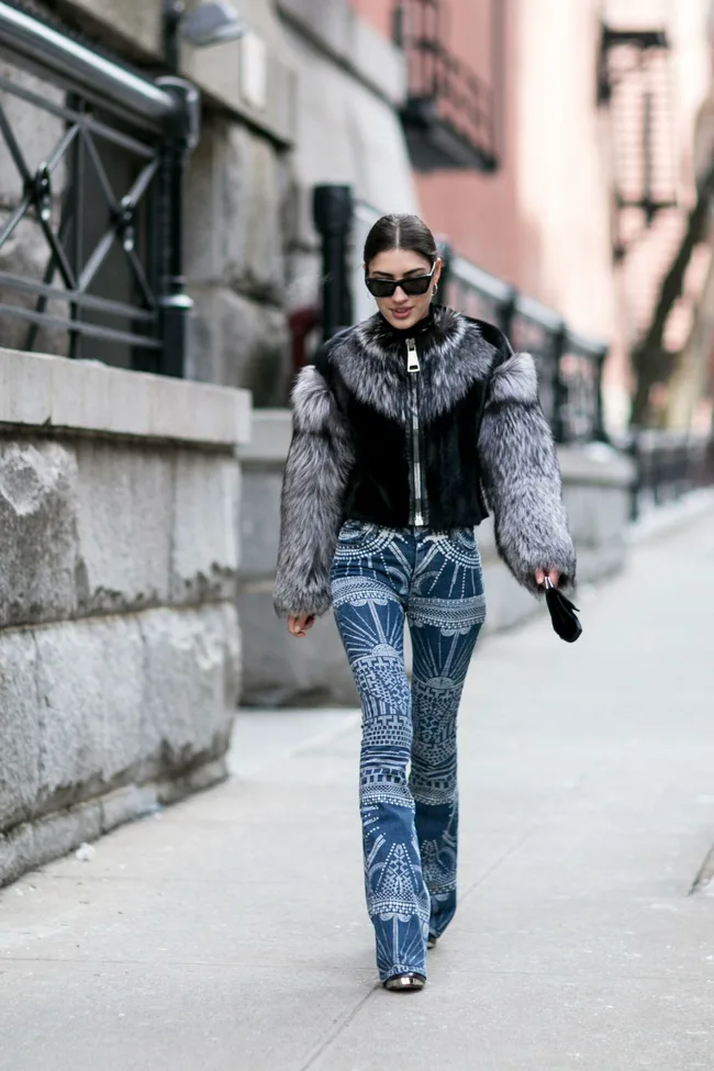 Woman in a fur jacket and patterned jeans walking down a city street, wearing sunglasses.