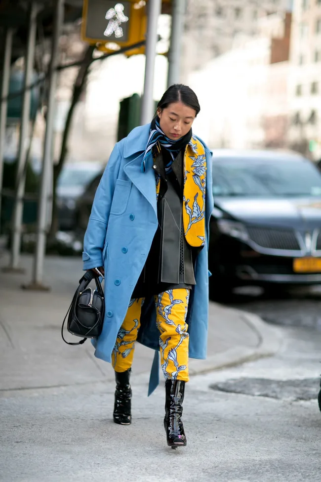 A person walking on a city street wearing a blue coat, patterned yellow pants, and carrying a black handbag.