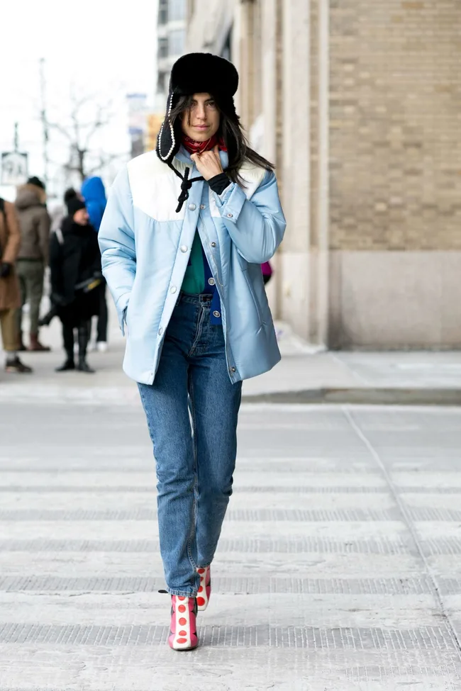 Person in blue jacket, jeans, and red spotted boots, wearing a black fur hat and red scarf, walking on a city street.