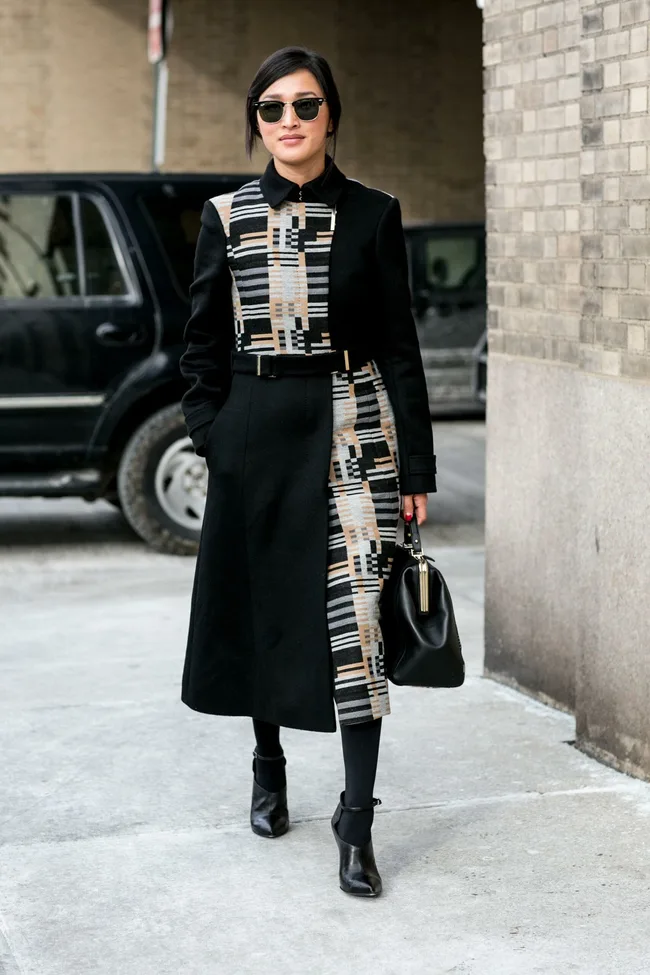 "Person in a stylish black coat with geometric patterns, wearing sunglasses and carrying a black bag, walking outdoors."