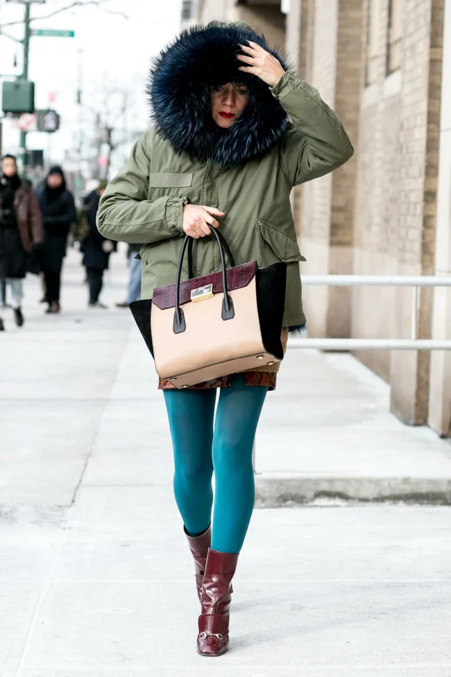 Person in a fur-hooded coat, blue tights, and maroon boots carrying a stylish handbag on a city sidewalk.