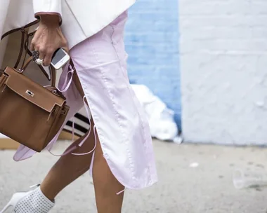 Person in white coat and pink pants walks down a street holding a brown handbag and a smartphone.