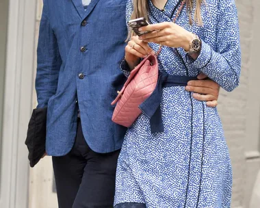 A person in a blue coat has their arm around another person in a blue patterned dress, who is holding a phone and a pink bag.