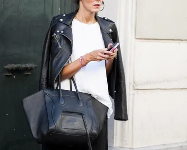 Woman in white shirt and black leather jacket holds phone and large black handbag.