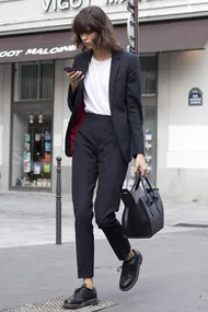 Woman in business attire, carrying a black bag, walking while looking at her phone on a city street.