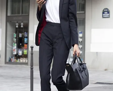 Woman in business attire, carrying a black bag, walking while looking at her phone on a city street.