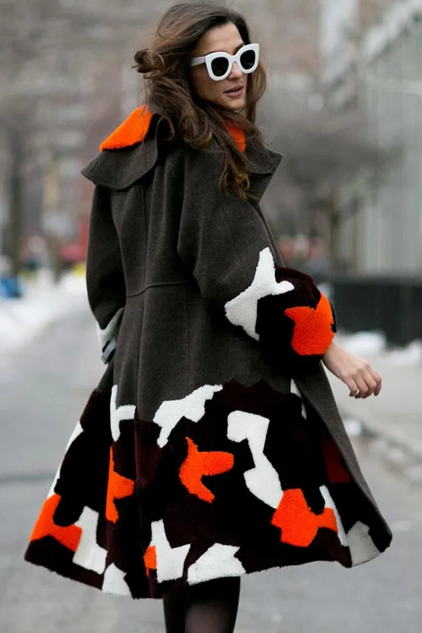 A woman in sunglasses wearing a colorful patterned coat with orange, white, and brown design, walking outdoors.