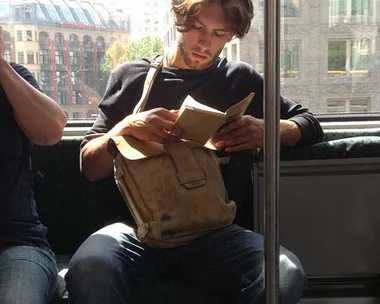 Man with long hair reading a book on a train, holding a tan satchel, with cityscape in the background.