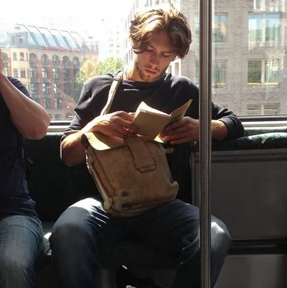 Man with long hair reading a book on a train, holding a tan satchel, with cityscape in the background.