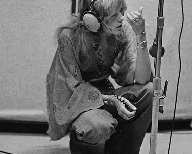 Person with headphones, kneeling near a mic stand in a recording studio, wearing flared sleeves. Black and white photo.