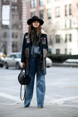 A woman wearing a large hat, fur scarf, flared jeans, and carrying a black handbag crosses an urban street.