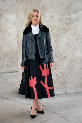 Woman in a leather jacket and black dress with red abstract print, standing against a textured concrete wall.