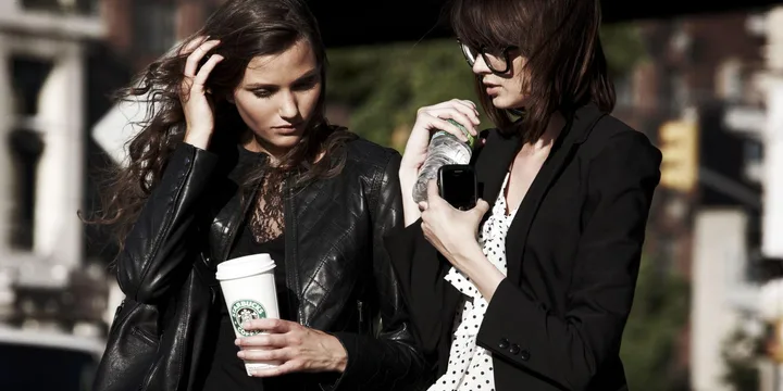 Two women in black jackets standing outdoors, one holding a Starbucks cup and the other a water bottle and phone.