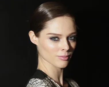 Woman with slicked-back hair and smoky eye makeup poses against a dark background.