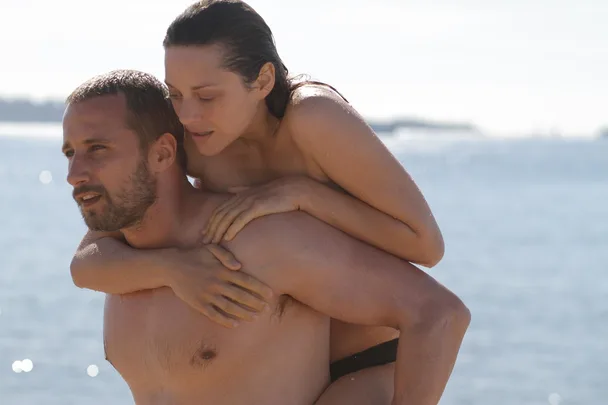 A shirtless man carries a woman piggyback on a beach, with the ocean background glistening in the sunlight.