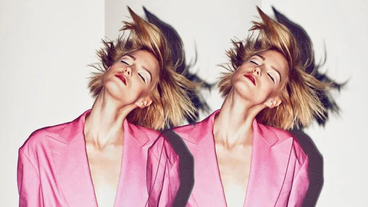 A woman in a bright pink blazer with short blonde hair is shown in a mirrored image, eyes closed and head tilted back.