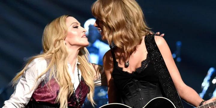 Madonna and Taylor Swift performing on stage, dressed in coordinating black and purple outfits, sharing a microphone and smiling.