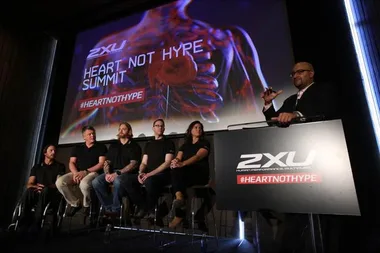 "Six people on stage at Heart Not Hype Summit by 2XU, speaker at podium, anatomical heart on screen, #HEARTNOTHYPE."