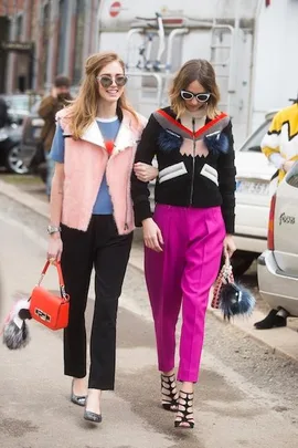 Two fashionable women walking on a street, wearing chic outfits with colorful accents and sunglasses, each carrying a trendy handbag.