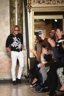 A designer wearing glasses, black shirt, and white pants is applauded by a seated audience at a fashion event.