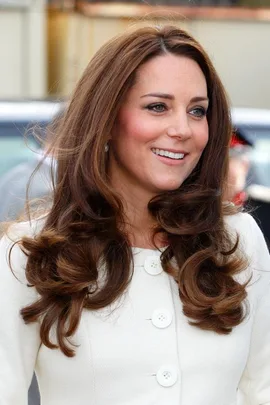 Kate Middleton with long wavy hair, wearing a white coat, smiling outdoors.