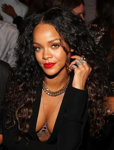 A person with long curly hair, wearing a black outfit, gold necklace, and red lipstick at a social event.