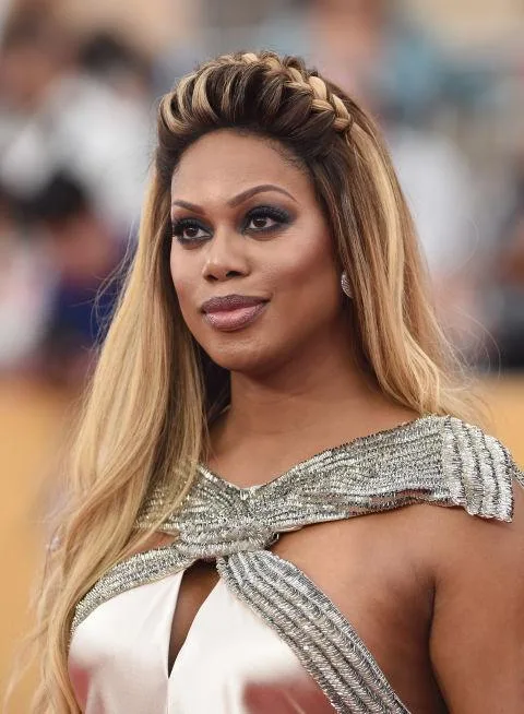 A person with long, ombre blonde hair and a braided crown, wearing a silver dress with metallic accents on a red carpet.