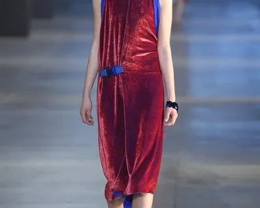 Model walks runway in red velvet halter dress with blue accents and heels during fashion show on concrete floor.