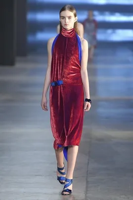 Model walks runway in red velvet halter dress with blue accents and heels during fashion show on concrete floor.