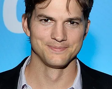 Man with short brown hair in a suit and open-collar shirt, standing against a blue background.