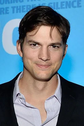 Man with short brown hair in a suit and open-collar shirt, standing against a blue background.