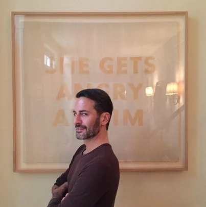 Man posing with a framed art piece behind him. The text on the art reads "SHE GETS ANGRY AT HIM."