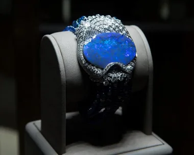 A luxurious bracelet featuring a large, vibrant blue gemstone set among sparkling diamonds on a cushioned display stand.