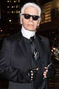 Karl Lagerfeld in a black suit, white shirt, black tie with a brooch, black sunglasses, and fingerless gloves, holding a phone.