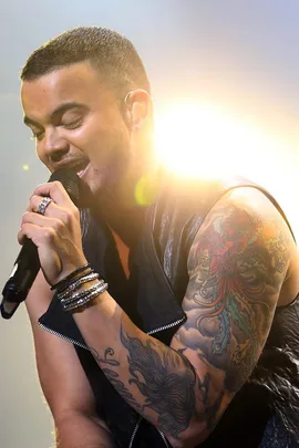 Singer with short hair and a tattooed arm performs on stage, holding a microphone, with bright light behind.