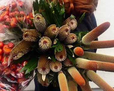 A person with red hair holds a large bouquet of unique, spiky flowers with green leaves and additional bouquets wrapped in plastic.