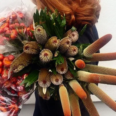 A person with red hair holds a large bouquet of unique, spiky flowers with green leaves and additional bouquets wrapped in plastic.