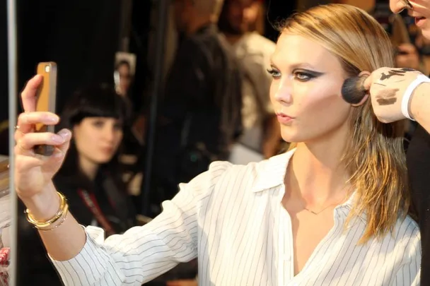 A woman taking a selfie while getting her makeup done, wearing a striped blouse.