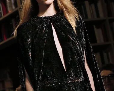 A model with long hair wears a dark, intricately patterned outfit on a catwalk, with bookshelves in the background.