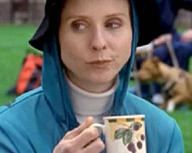 Miranda from "Sex and the City" wearing a blue hoodie and black hat, holding a white mug with a fruit design in a park.