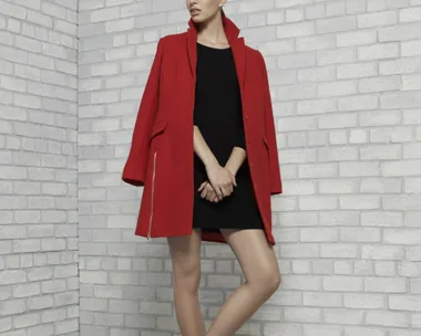 A woman in a red coat and black dress stands by a white brick wall, wearing white shoes with tassels.