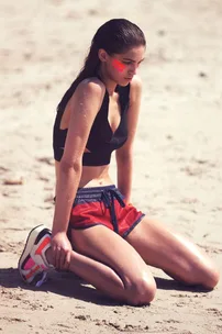 A woman in a black sports bra and red shorts kneeling on a sandy beach, with red paint stripes on her cheeks.