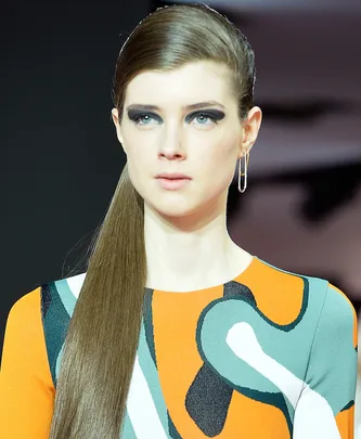 Model with sleek hair and hoop earrings on runway, wearing colorful abstract-patterned outfit at Paris Fashion Week.